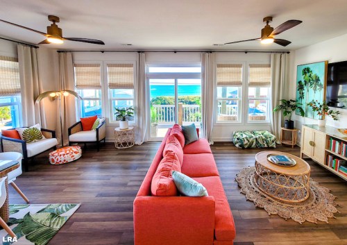 Bright living room with a coral sofa, tropical decor, and large windows showcasing a view of the ocean.