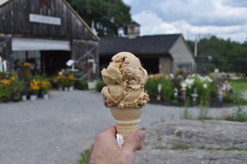 A hand holds a large cone of ice cream in front of a rustic building and a garden filled with flowers.