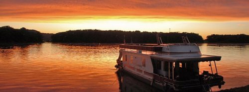 A houseboat anchored on calm waters during a vibrant sunset, with colorful skies reflecting on the surface.