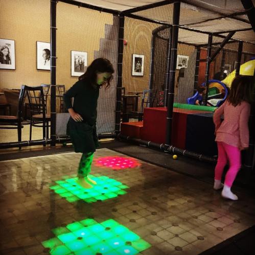 Two children play on a colorful, illuminated floor in a play area, with a netted enclosure and playful decor in the background.