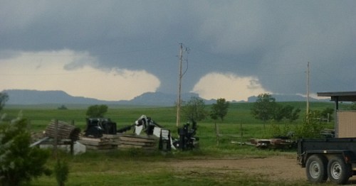 Dark storm clouds loom over a green landscape, with distant mountains and a rural scene featuring equipment and a trailer.