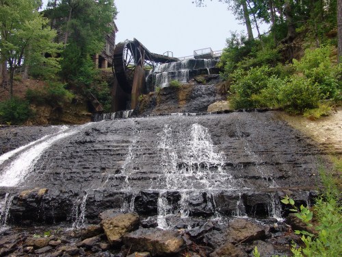 A cascading waterfall flows over rocky terrain, with a waterwheel and lush greenery in the background.