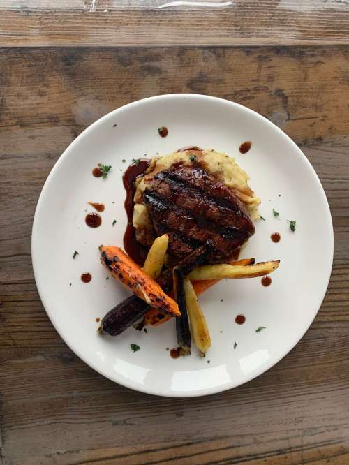 Grilled steak on a bed of mashed potatoes, garnished with colorful roasted carrots and drizzled with sauce.