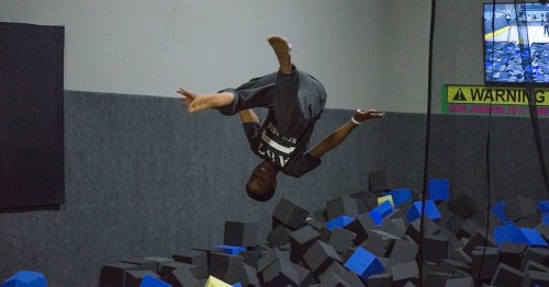 A child performs a backflip above a foam pit in a trampoline park, with a safety warning sign in the background.