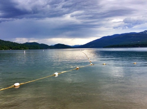 A calm lake with a buoyed rope stretching across the water, surrounded by mountains under a cloudy sky.