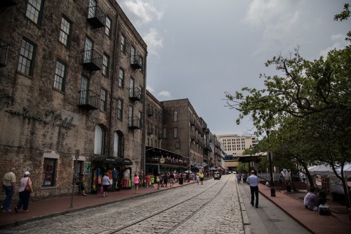 Historic street scene with brick buildings, shops, and people walking along a cobblestone path under a cloudy sky.