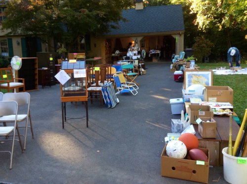 A yard sale with various items for sale, including furniture, sports equipment, and boxes, set in a driveway.