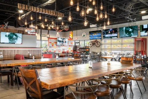 A spacious restaurant interior with wooden tables, hanging lights, and multiple TVs displaying sports.