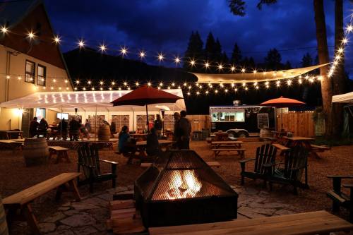 A cozy outdoor gathering space at night, featuring string lights, a fire pit, and people enjoying food and drinks.