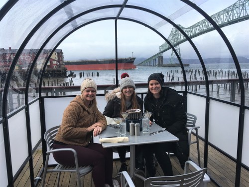 Three women sit at a table in a glass-enclosed patio by the water, wearing winter hats, with a ship and bridge in the background.