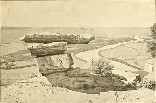 A large, flat rock balanced on top of stacked stones, overlooking a winding river and lush landscape.