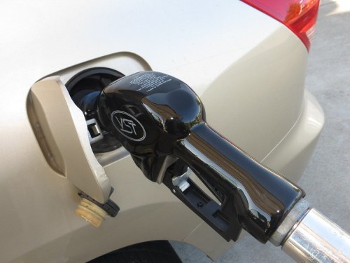 A close-up of a fuel nozzle inserted into a car's gas tank, with the vehicle's rear visible in the background.