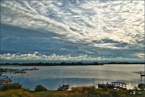 A serene lake scene with boats, lush greenery, and a dramatic sky filled with clouds reflecting on the water.