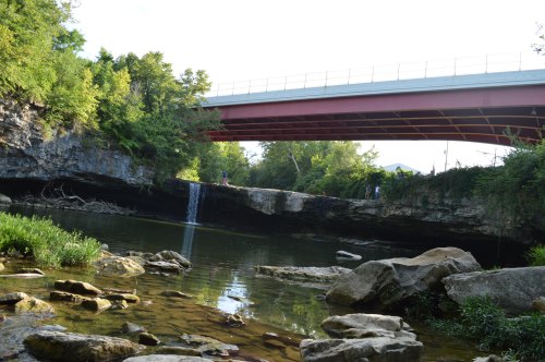 A serene landscape featuring a waterfall under a bridge, surrounded by lush greenery and rocky terrain.