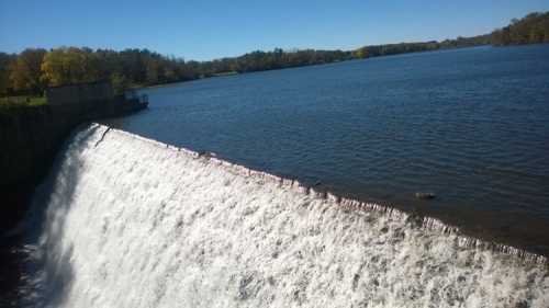 A waterfall cascading over a dam into a calm lake, surrounded by trees and clear blue skies.