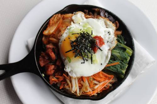 A sizzling skillet filled with colorful vegetables, kimchi, and a fried egg, garnished with green onions and seaweed.