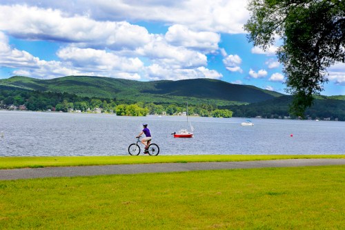 A person rides a bicycle along a lakeside path with boats on the water and mountains in the background under a blue sky.