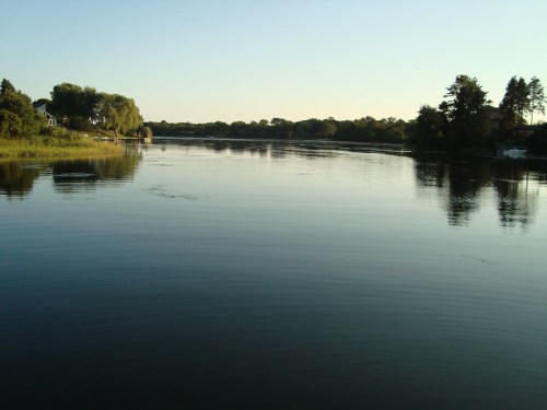 A calm river reflects the clear sky, surrounded by trees and houses on the banks.
