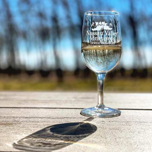 A wine glass with "Nassau Valley Vineyards" etched on it, casting a shadow on a wooden table outdoors.