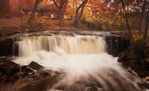 A serene waterfall flows through a forest with autumn foliage, while a cozy cabin is visible in the background.