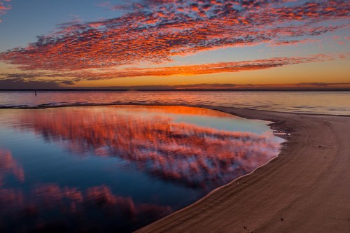A serene beach at sunset, with vibrant clouds reflecting in calm water and a sandy shore.