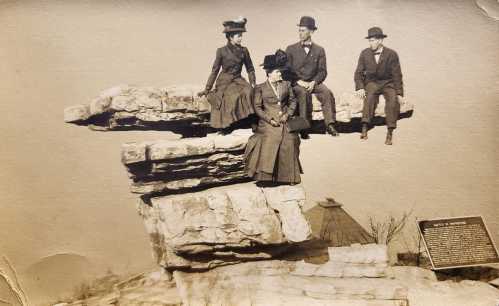 Four people in vintage clothing sit on a large rock formation, with a plaque and a distant landscape in the background.