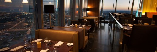 Elegant restaurant interior with city skyline views at sunset, featuring tables set for dining.