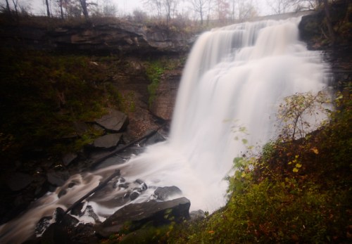 A serene waterfall cascading over rocky terrain, surrounded by autumn foliage and a misty atmosphere.