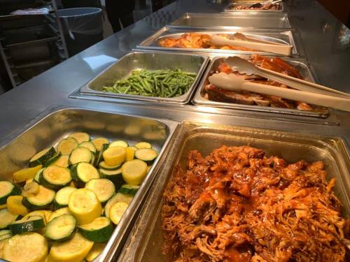 A buffet spread featuring green beans, zucchini, pulled chicken in sauce, and other dishes in metal trays.