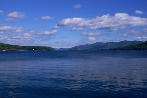 A serene lake surrounded by green hills under a blue sky with fluffy clouds, reflecting on the water's surface.
