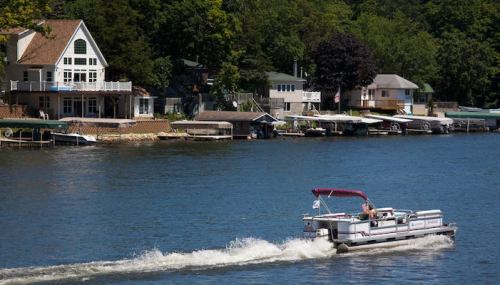 A boat glides across a calm lake, with houses and trees lining the shore in the background.