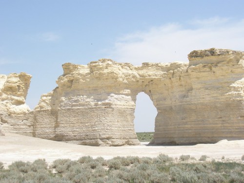 A large, natural rock formation with an arch, surrounded by sparse vegetation under a clear blue sky.