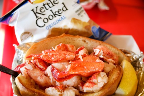A lobster roll filled with chunks of lobster meat, served with a lemon wedge and a bag of kettle-cooked chips.