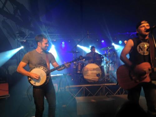 A band performs on stage with a banjo player and a guitarist, illuminated by colorful stage lights.