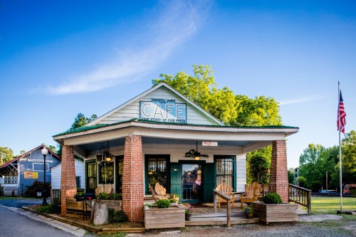 A charming café with a porch, surrounded by greenery and an American flag, under a clear blue sky.