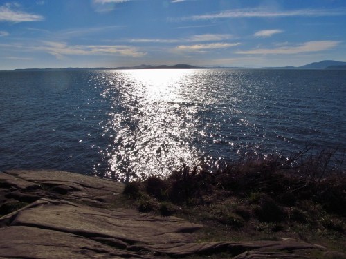 A serene view of shimmering water under a bright sky, with rocky shoreline in the foreground and distant islands.