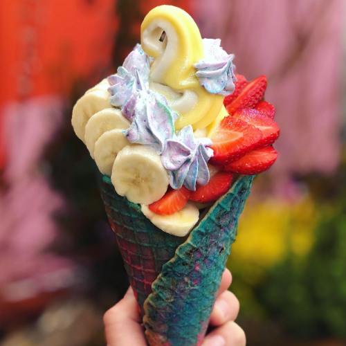 A colorful ice cream cone topped with swirls of soft serve, strawberries, bananas, and vibrant whipped cream.
