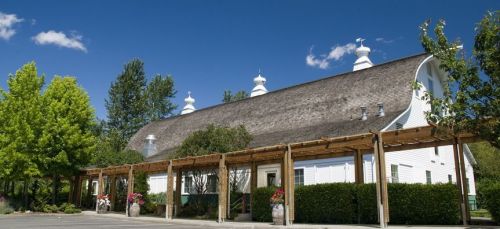 A large white barn with a curved roof, surrounded by green trees and a wooden trellis with flowers. Blue sky above.
