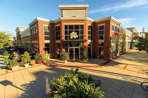 A modern brick building with large windows, featuring shops and outdoor seating, surrounded by planters and trees.