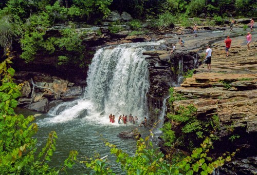 A scenic waterfall surrounded by lush greenery, with people swimming and enjoying the area.