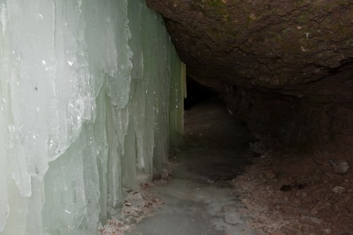 A narrow cave passage with icy walls and a frozen floor, illuminated by faint light.