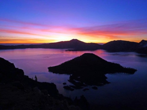 A vibrant sunset over a lake, with silhouettes of mountains and an island in the foreground.