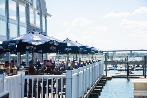 Outdoor dining area with umbrellas overlooking a marina, featuring people enjoying the sunny day by the water.