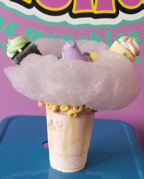 A colorful dessert in a cup topped with cotton candy, cupcakes, and cereal pieces against a purple background.