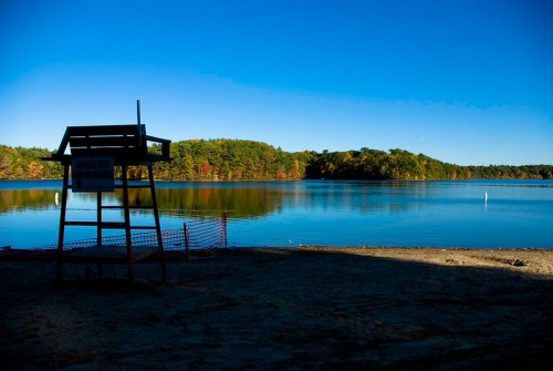 A calm lake surrounded by trees in autumn colors, with a lifeguard stand and blue sky above.