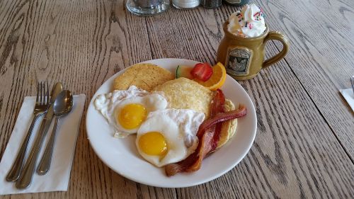 A plate of breakfast with two fried eggs, bacon, pancakes, and fruit, alongside a mug of whipped cream topped drink.