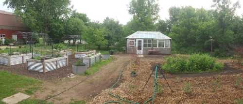 A garden with raised beds, a greenhouse, and lush greenery in a rural setting.