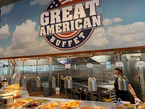A buffet with a "Great American Buffet" sign, featuring various food dishes and a staff member wearing a mask.