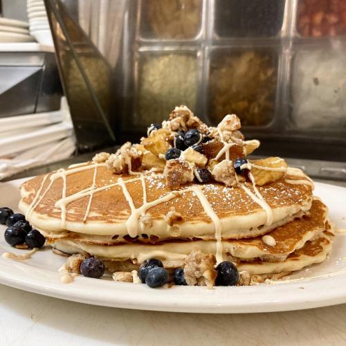 A stack of pancakes topped with blueberries, crumbled nuts, and a drizzle of icing on a white plate.