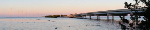 A panoramic view of a bridge over calm water, with power lines and trees in the foreground during sunset.